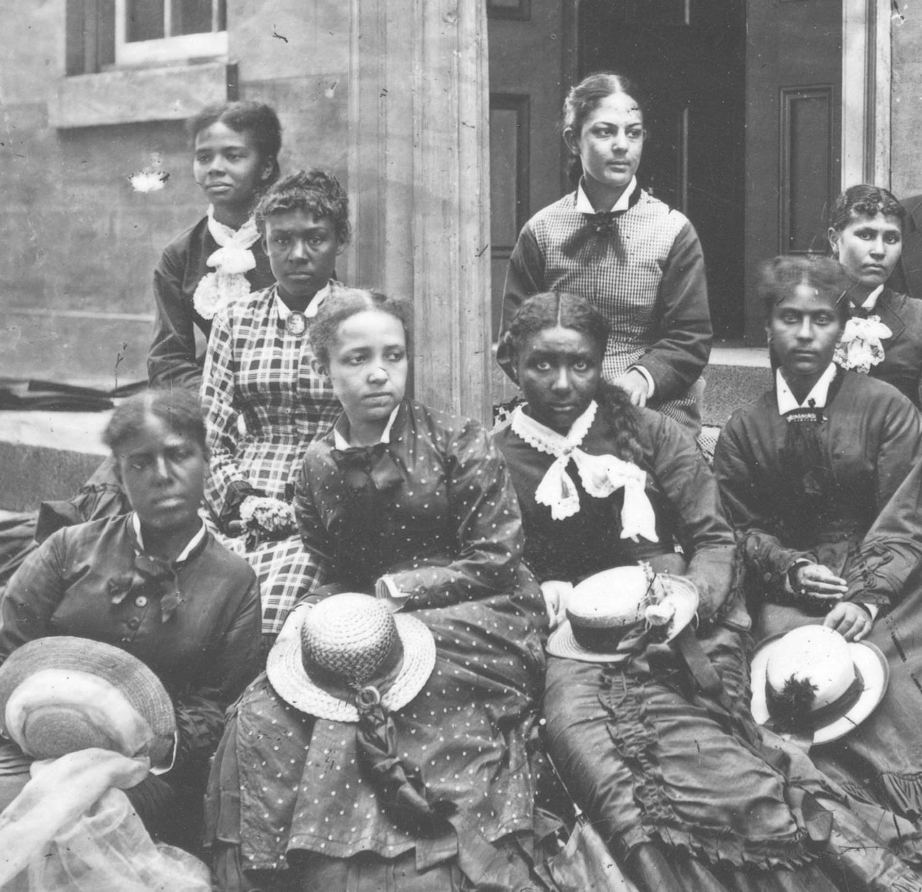 Women in Victorian dresses hold hats and look solemn outside a stone building.
