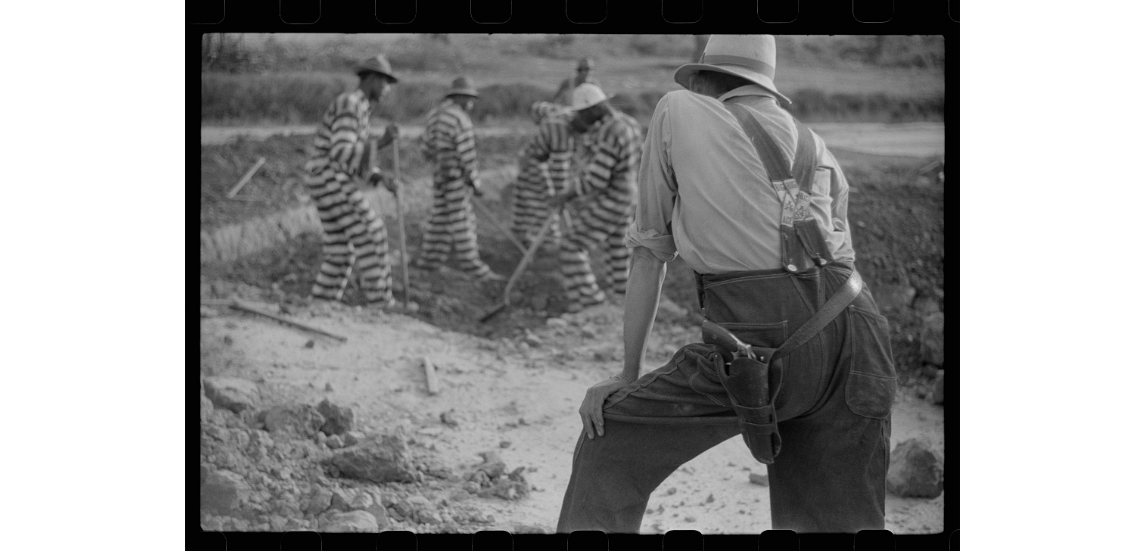 Black and white photo of a man in overalls with a gun on his hip watching Black men in striped prison suits work in a field