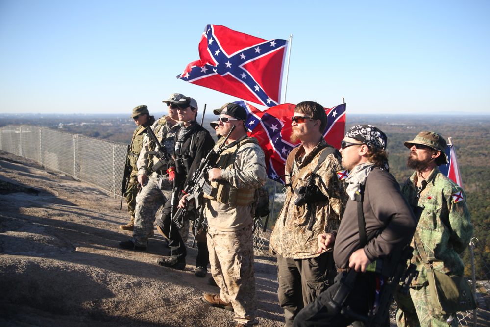 A group of individuals wearing camouflage and sunglasses and carrying rifles and confederate flags stand on a hillside