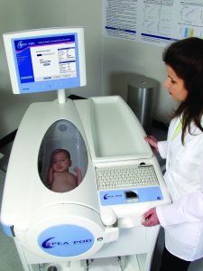 A baby can be seen through an observation window on a large machine in which the baby is enclosed. A healthcare professional operates the machine to measure the baby's body fat percentage.