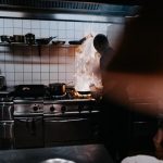 In a kitchen, flames are rising from a pan next to a person. 