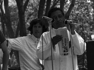 Photo of César Chávez holding a microphone and a folded newspaper