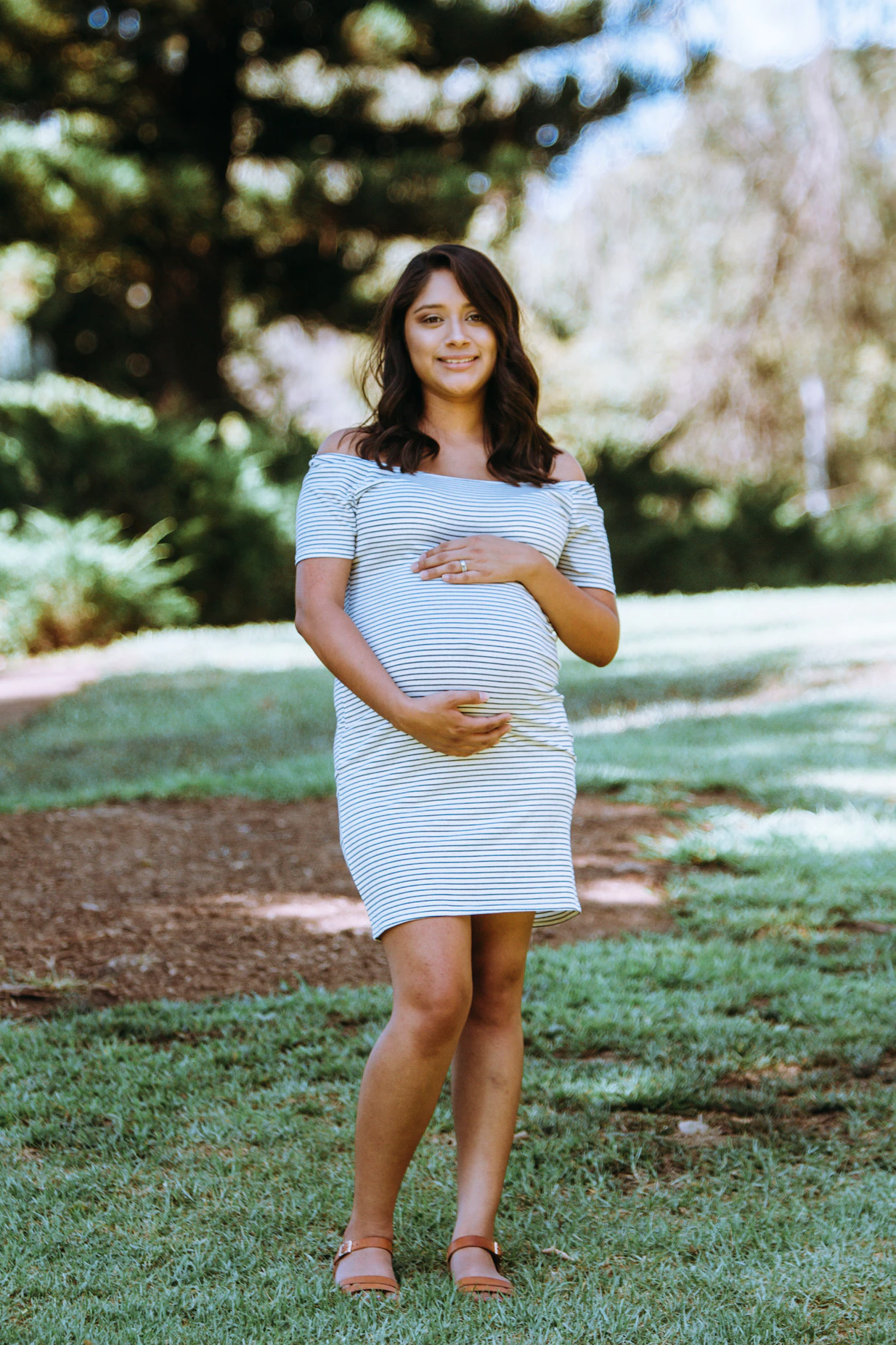 A pregnant woman with medium skin tone and long, dark hair stands outside with her arms encircling her belly.