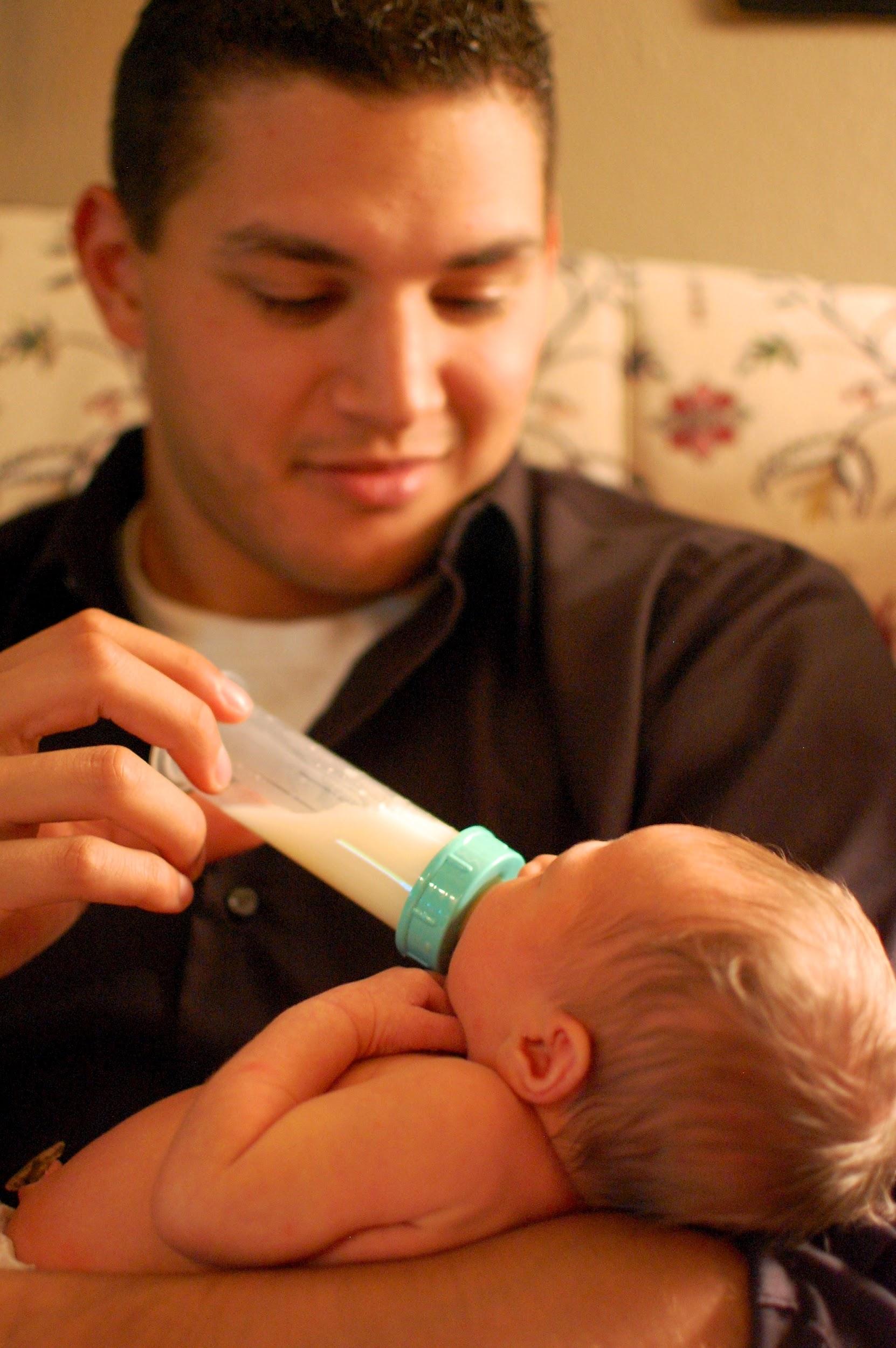 A father with medium skin tone and dark hair bottle-feeds a newborn infant. The father is smiling and gazing at the baby.